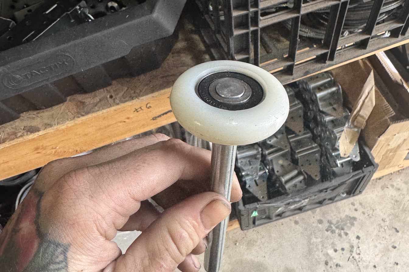 A nylon-tyred garage door roller held in one hand, with a crate of plain steel rollers on the shelf behind it